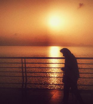 Silhouette of a person walking along a railing with a dramatic sunset over the water.