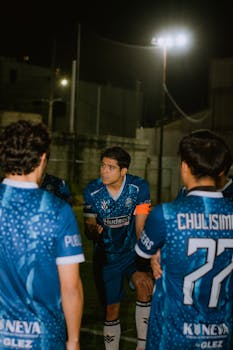 Amateur soccer team players huddle on the field under floodlights during a night match.
