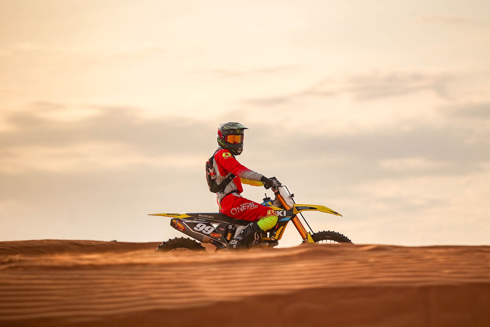 A motocross rider in red gear riding across desert dunes during a beautiful sunset.