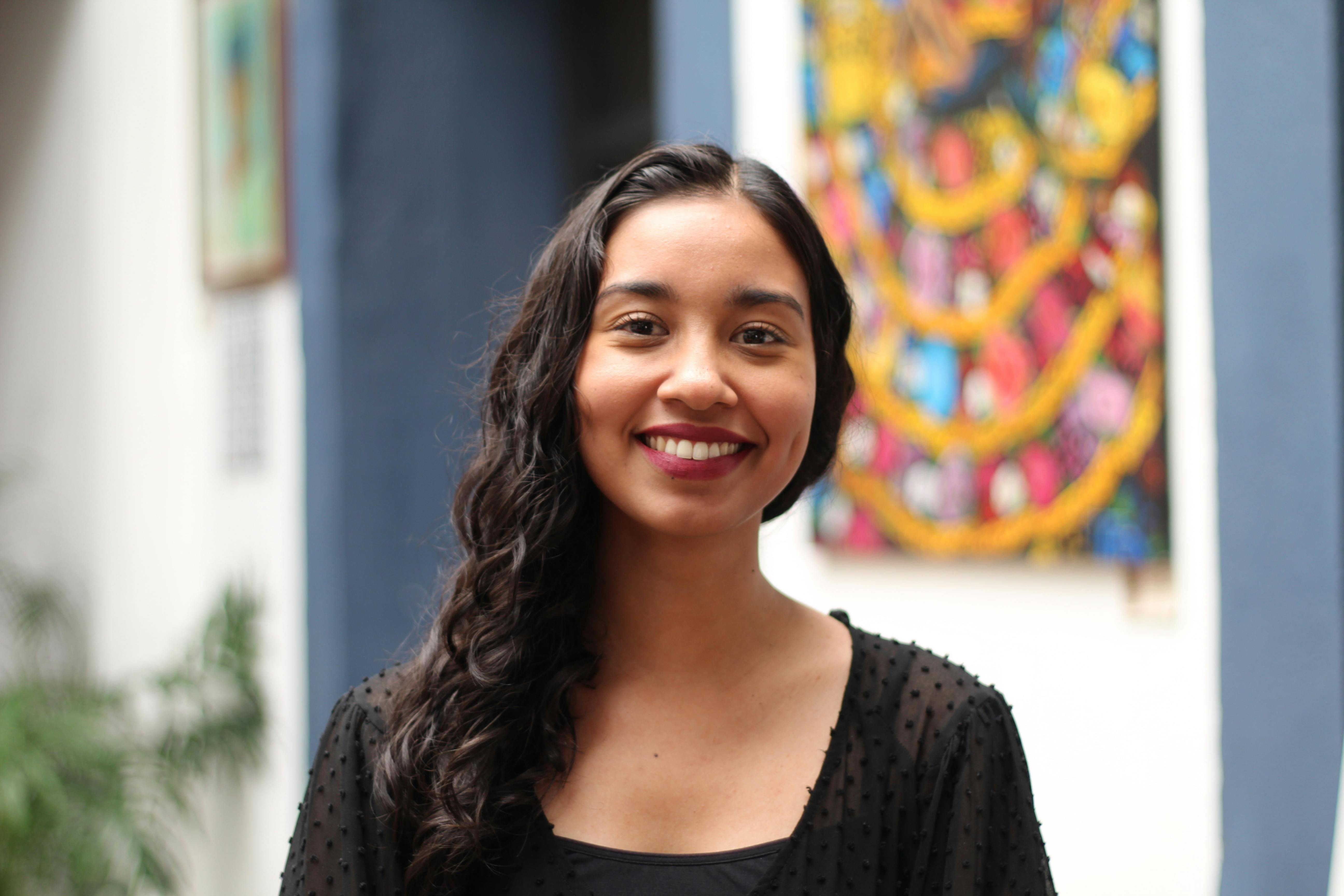 A woman smiles brightly in an artistic indoor setting with vibrant artwork in the background.