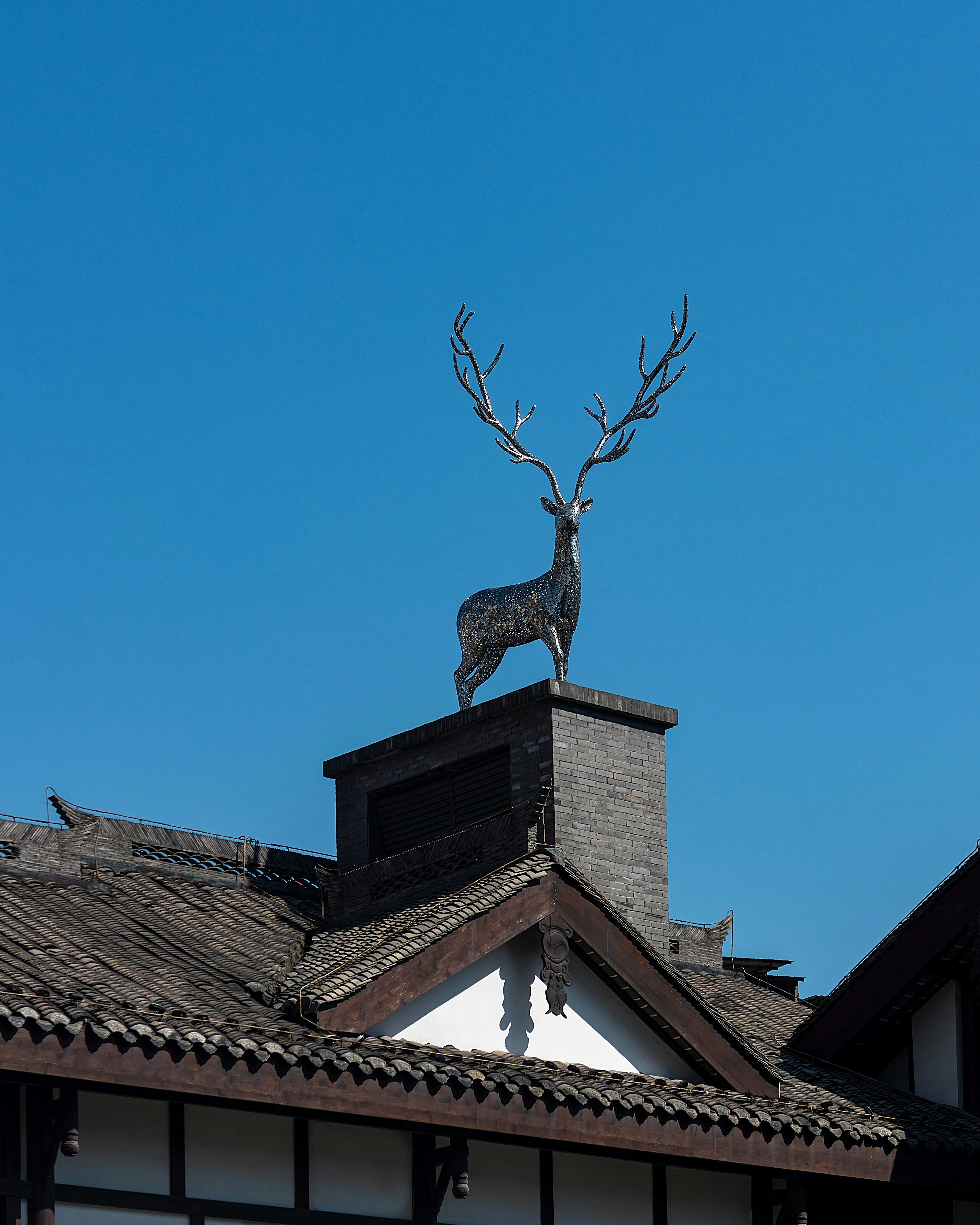 grátis Uma impressionante escultura de veado adorna um telhado tradicional contra um céu azul claro, exibindo arte arquitetônica. Foto profissional