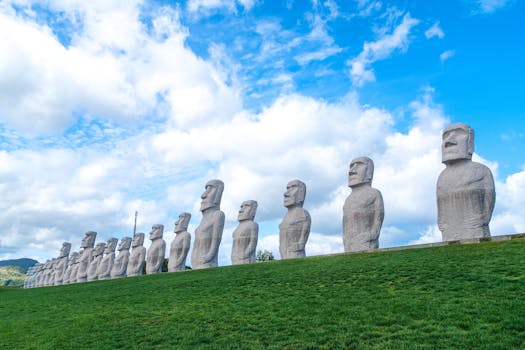 A striking alignment of Moai-inspired statues under a bright, cloudy sky on grass-covered hills.