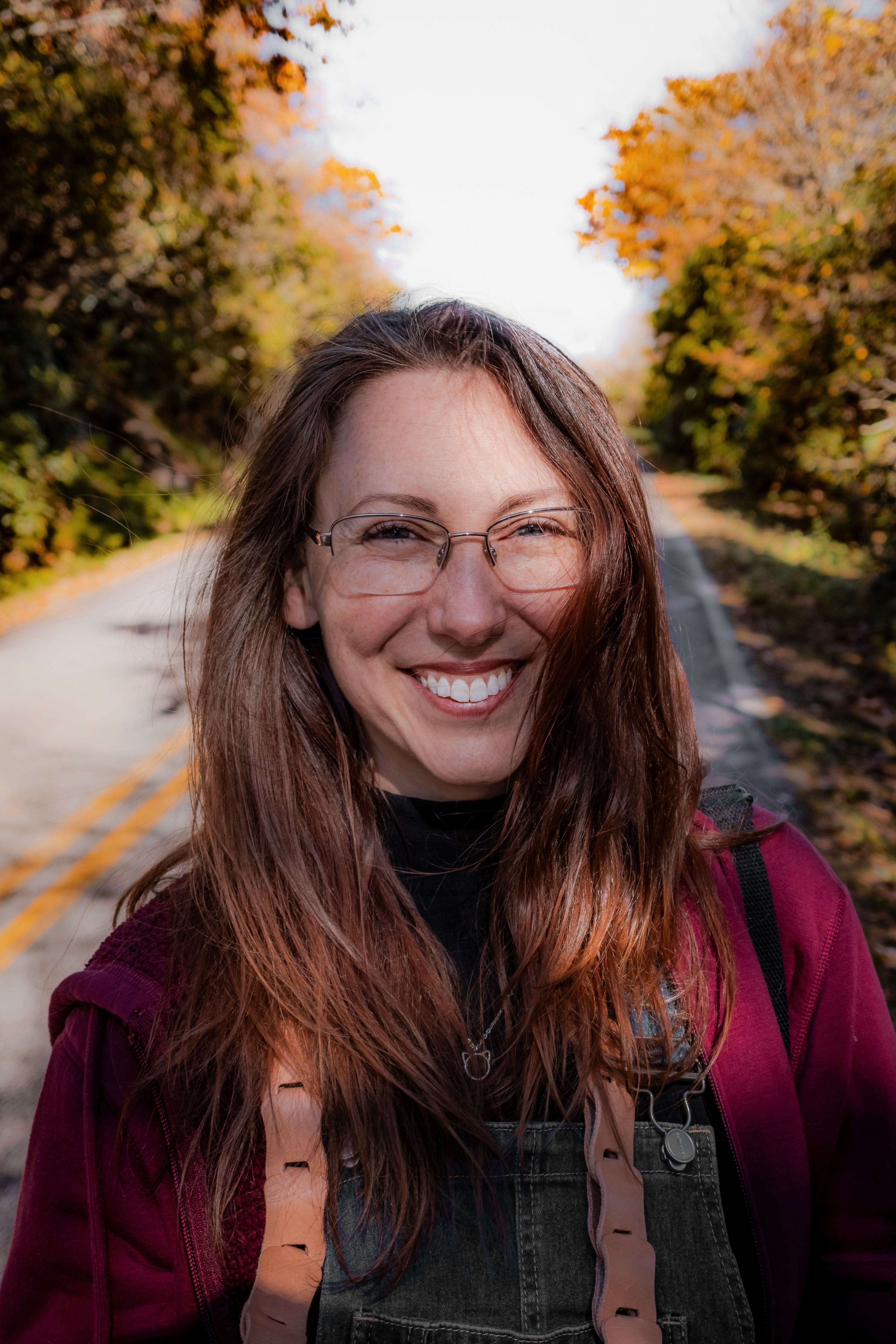 A joyful woman with glasses smiles on a rural road surrounded by autumn foliage.
