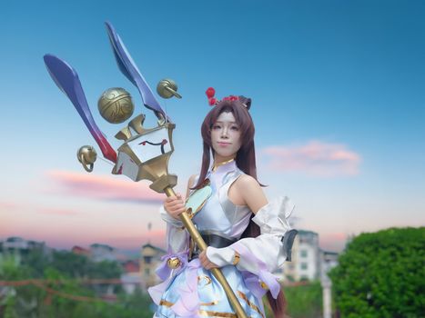 A cosplayer in a detailed fantasy outfit poses with a large ornate staff against a vibrant sky.