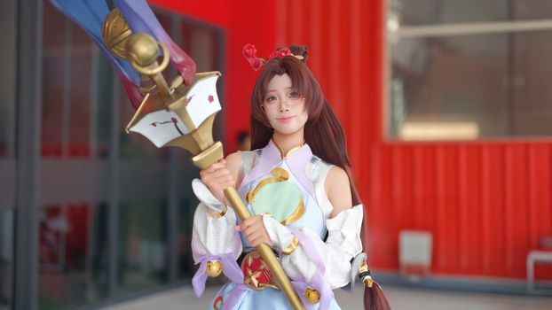 A cosplayer in a detailed costume poses with a large staff, showcasing colorful and intricate design outdoors.