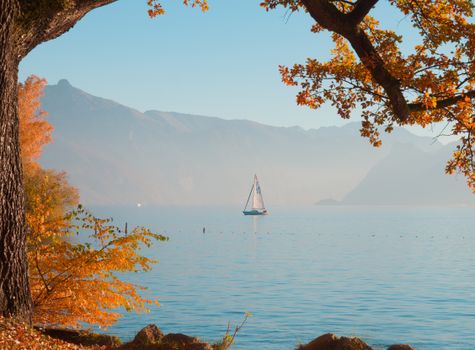A tranquil autumn view of a sailboat on a lake framed by colorful trees and distant mountains.