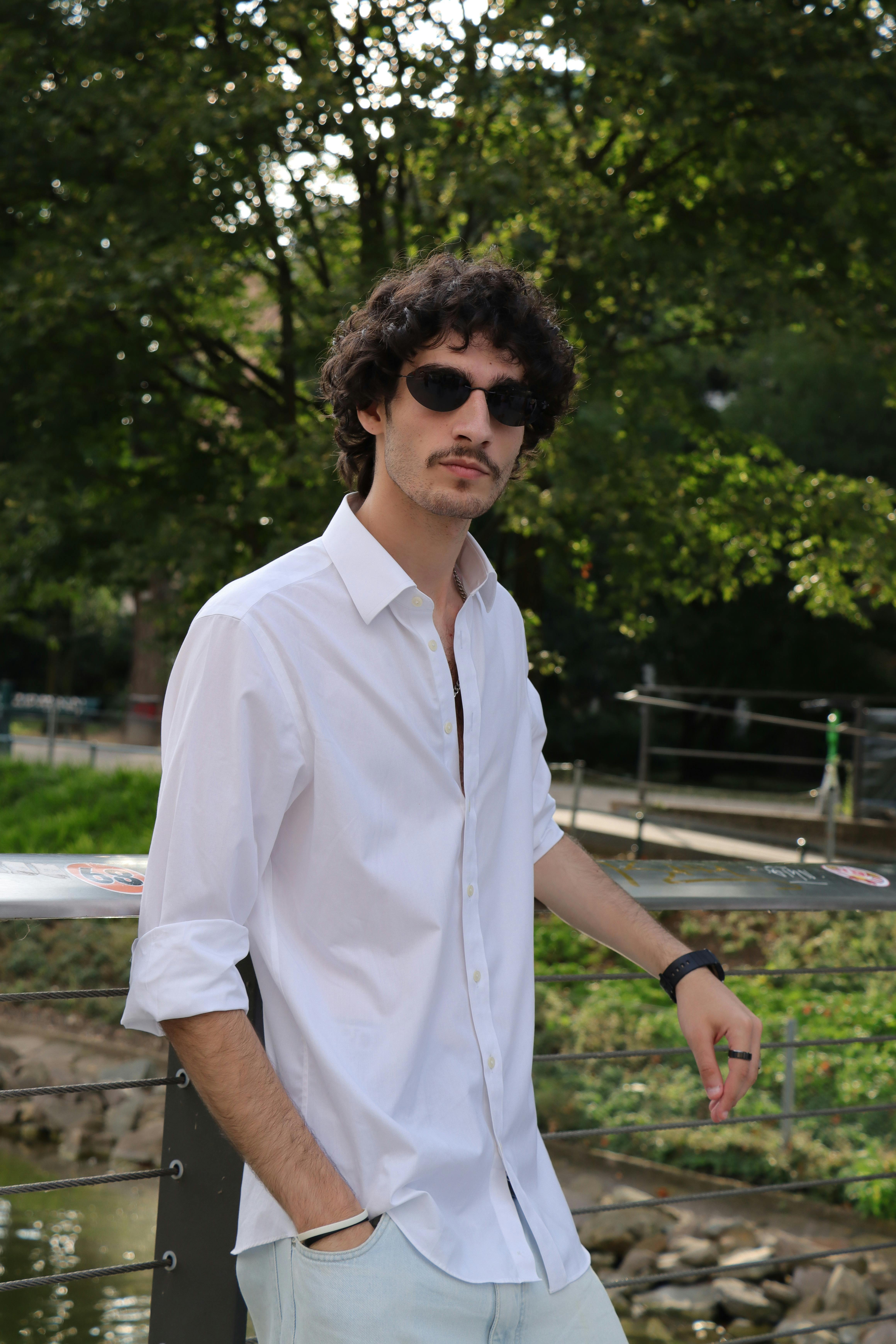 Young man in a white shirt and sunglasses posing confidently outdoors with greenery in the background.