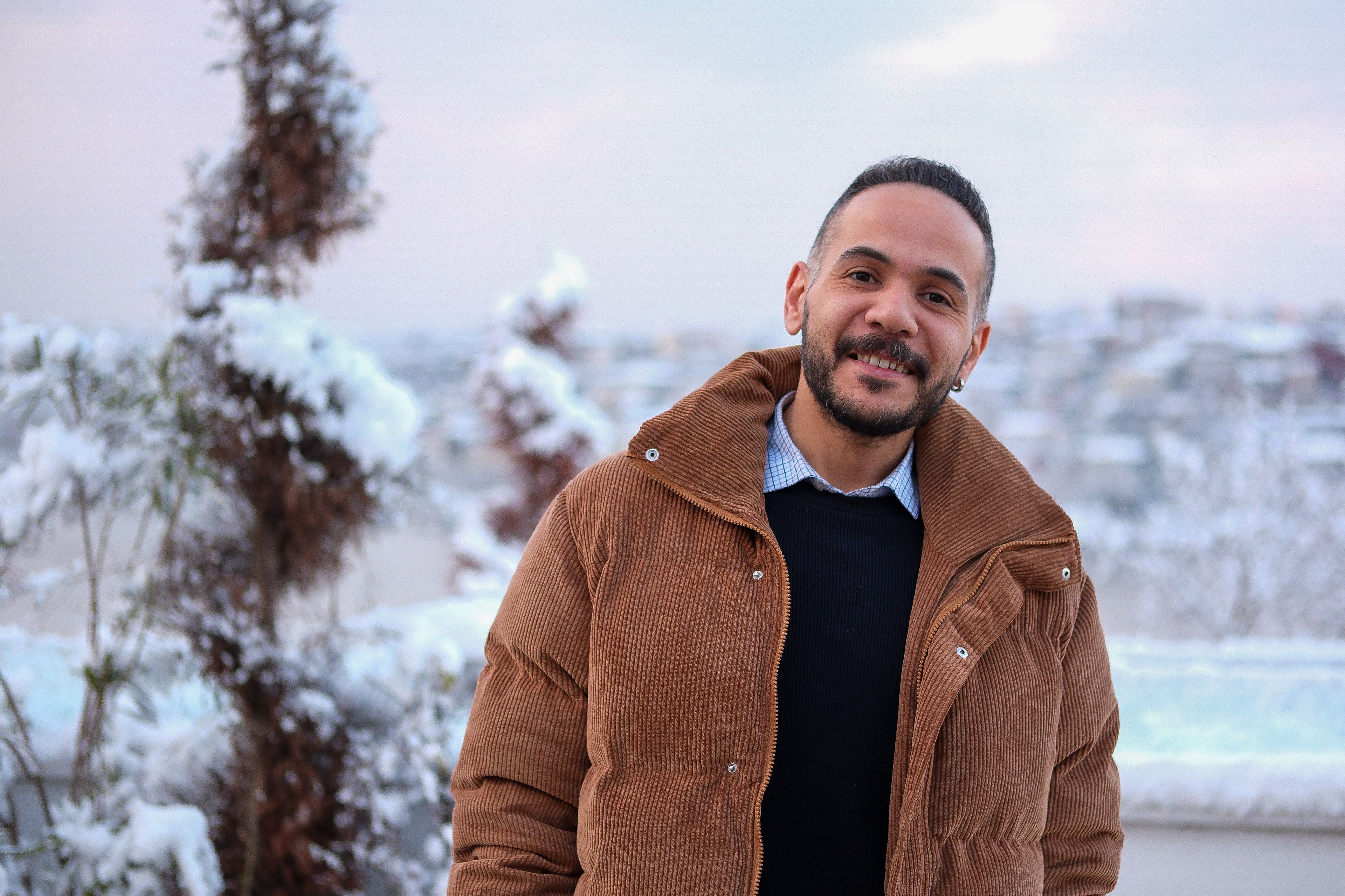 Cheerful man with a beard in a warm winter jacket standing outdoors with snow in the background.