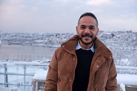 A man in a brown winter coat smiles outdoors against a snowy cityscape backdrop.