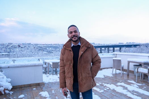 Smiling man on a snowy rooftop terrace with a scenic city view in winter.