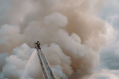 Free Firefighter on ladder extinguishing fire, enveloped by thick smoke in daylight. Stock Photo