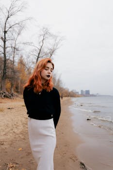 A young woman with red hair walks along a serene beach in autumn, dressed stylishly in a monochrome outfit.