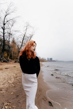A young woman with red hair stands on a sandy beach during autumn. Moody and serene scene.