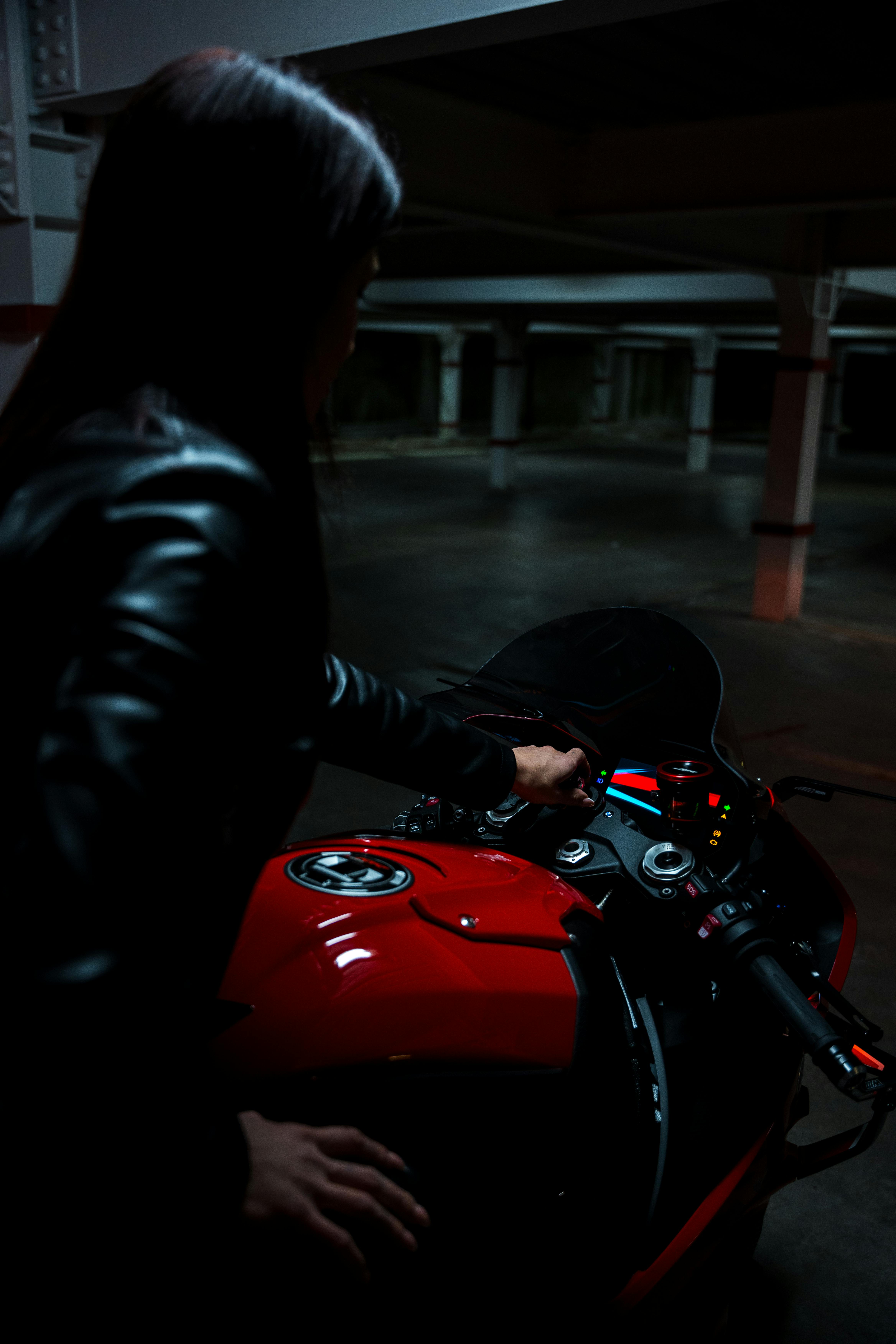 A woman in a black leather jacket on a red motorcycle in a dimly lit garage.