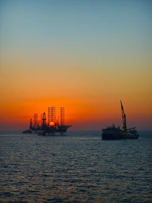 Free Dramatic view of an offshore oil rig and ship silhouetted against a vibrant sunset over the ocean. Stock Photo