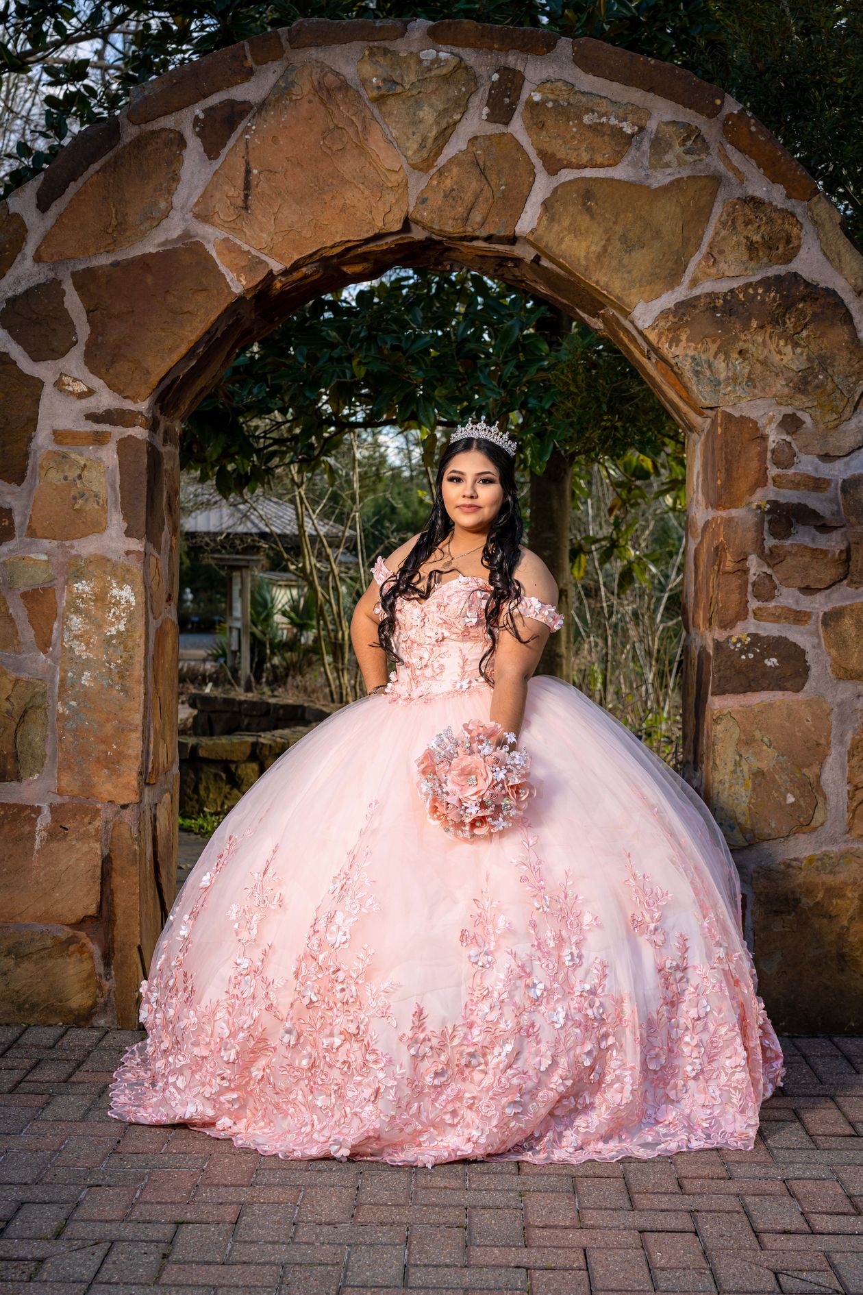 Young woman in a pink off-the-shoulder quinceañera ball gown under a stone arch