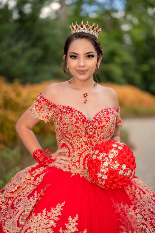 Free Elegant young woman in a vibrant red quinceañera gown with golden embroidery and crown, posing confidently outdoors. Stock Photo