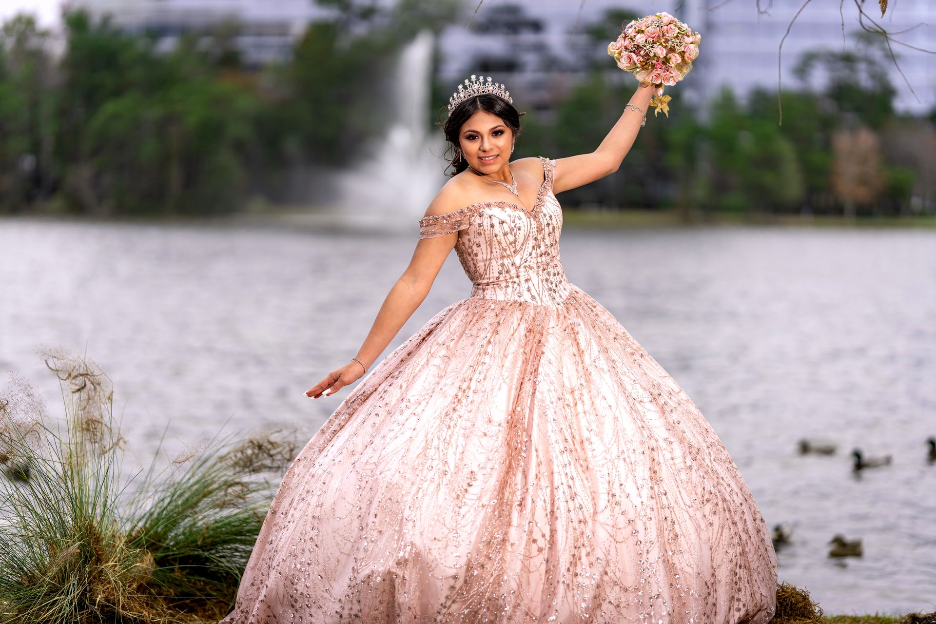 A young woman celebrates her quinceaƱera in a stunning gown by a scenic lake, holding a bouquet.