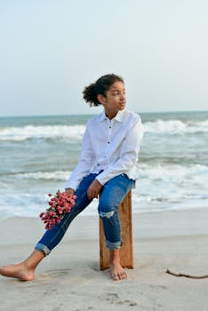 A young boy in a white shirt sits on a beach holding pink flowers, enjoying the ocean view.