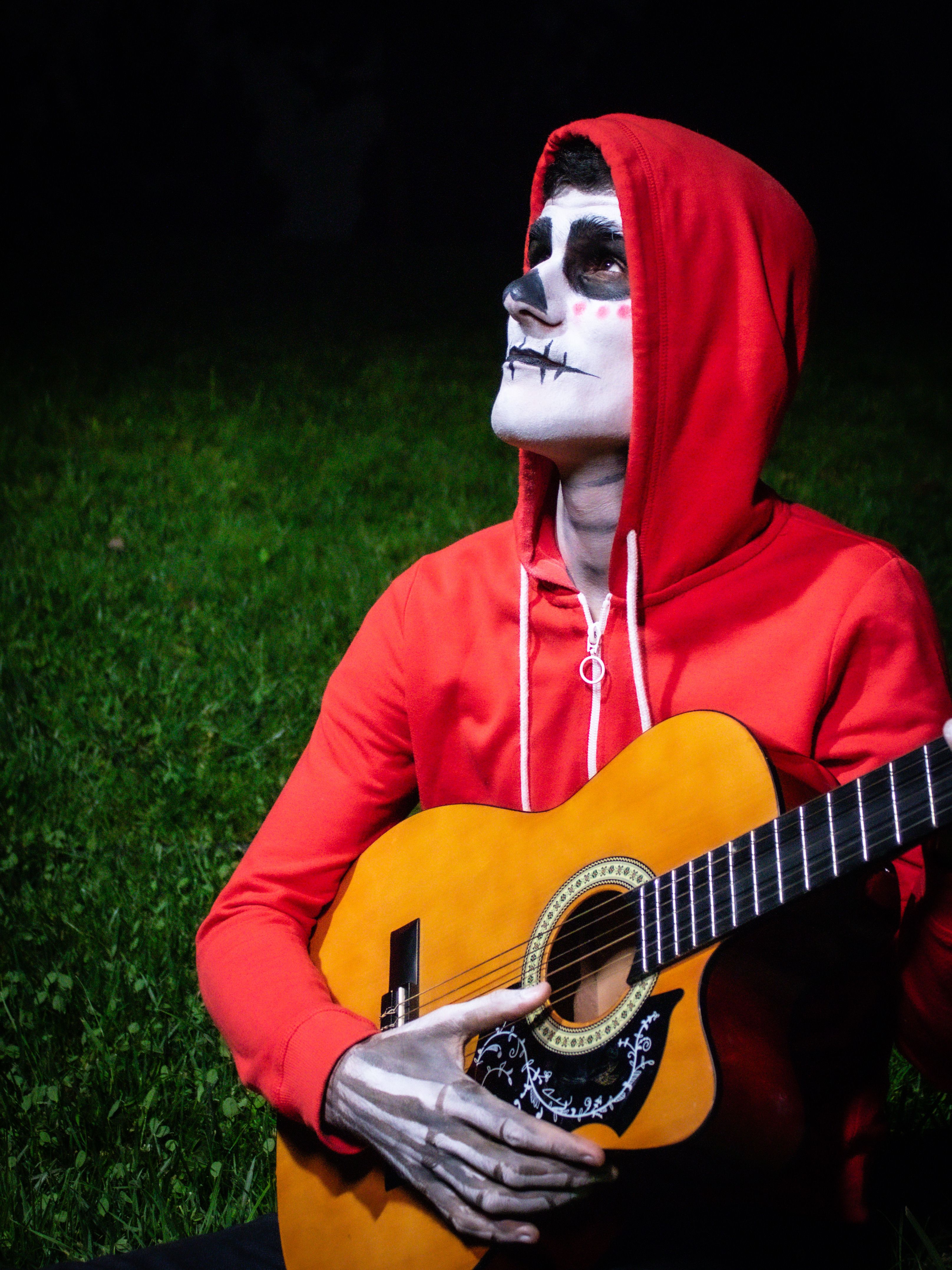 Person with skull makeup wearing red hoodie playing guitar outdoors at night.