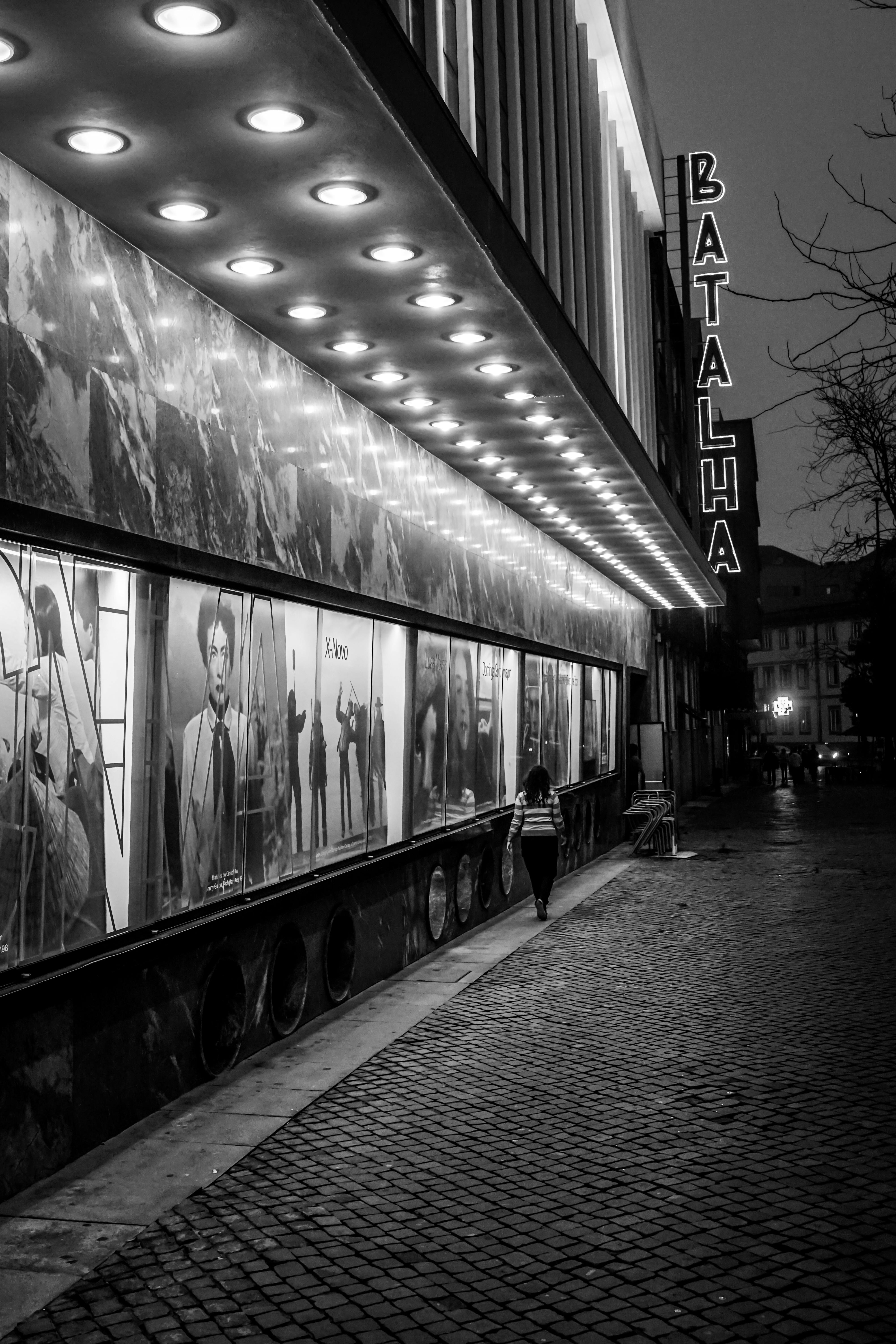 Free Black and white photo of a person walking past the illuminated Batalha cinema at night. Stock Photo
