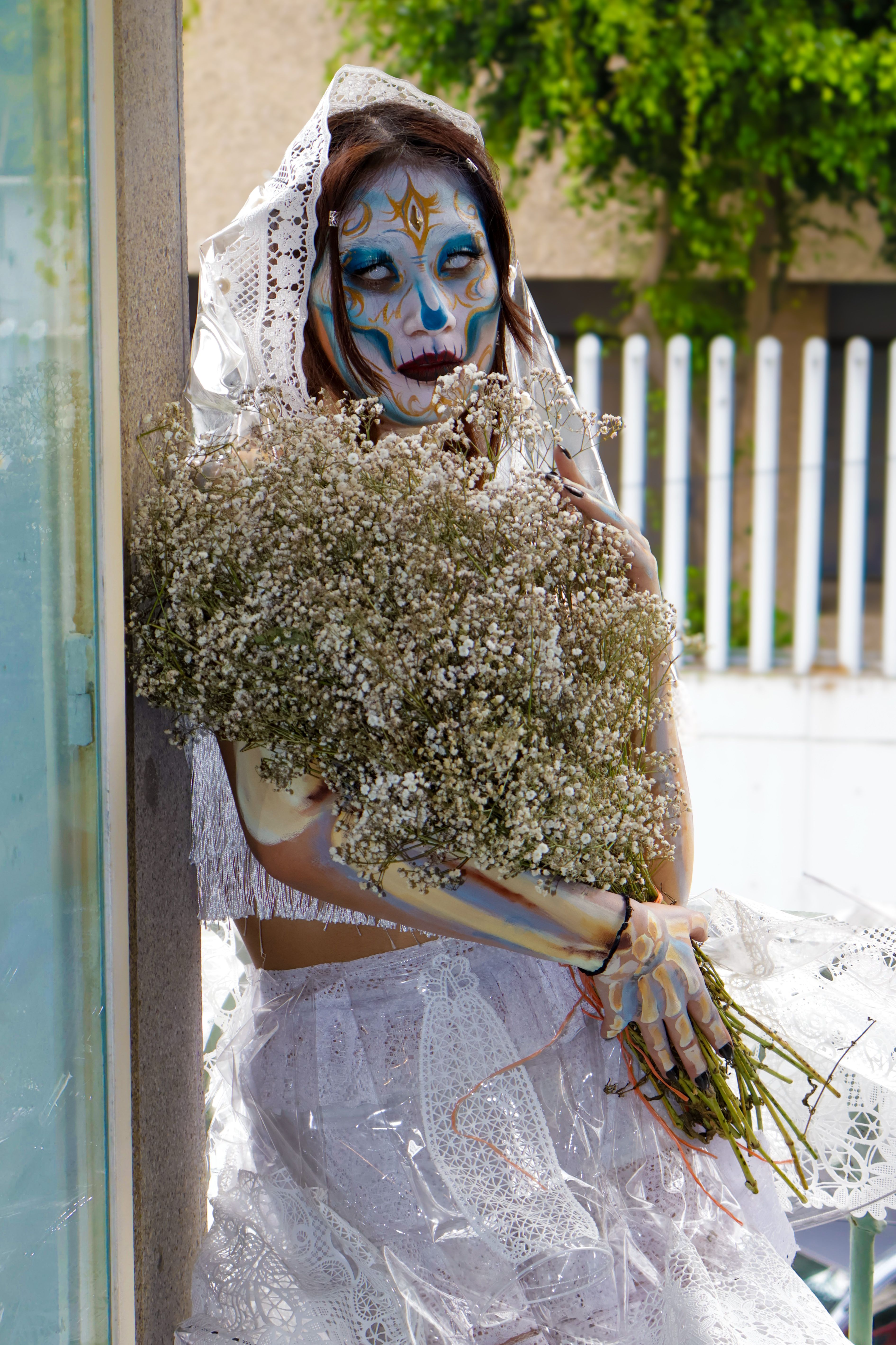 A Day of the Dead bride with intricate face paint holding a bouquet of baby's breath outdoors.