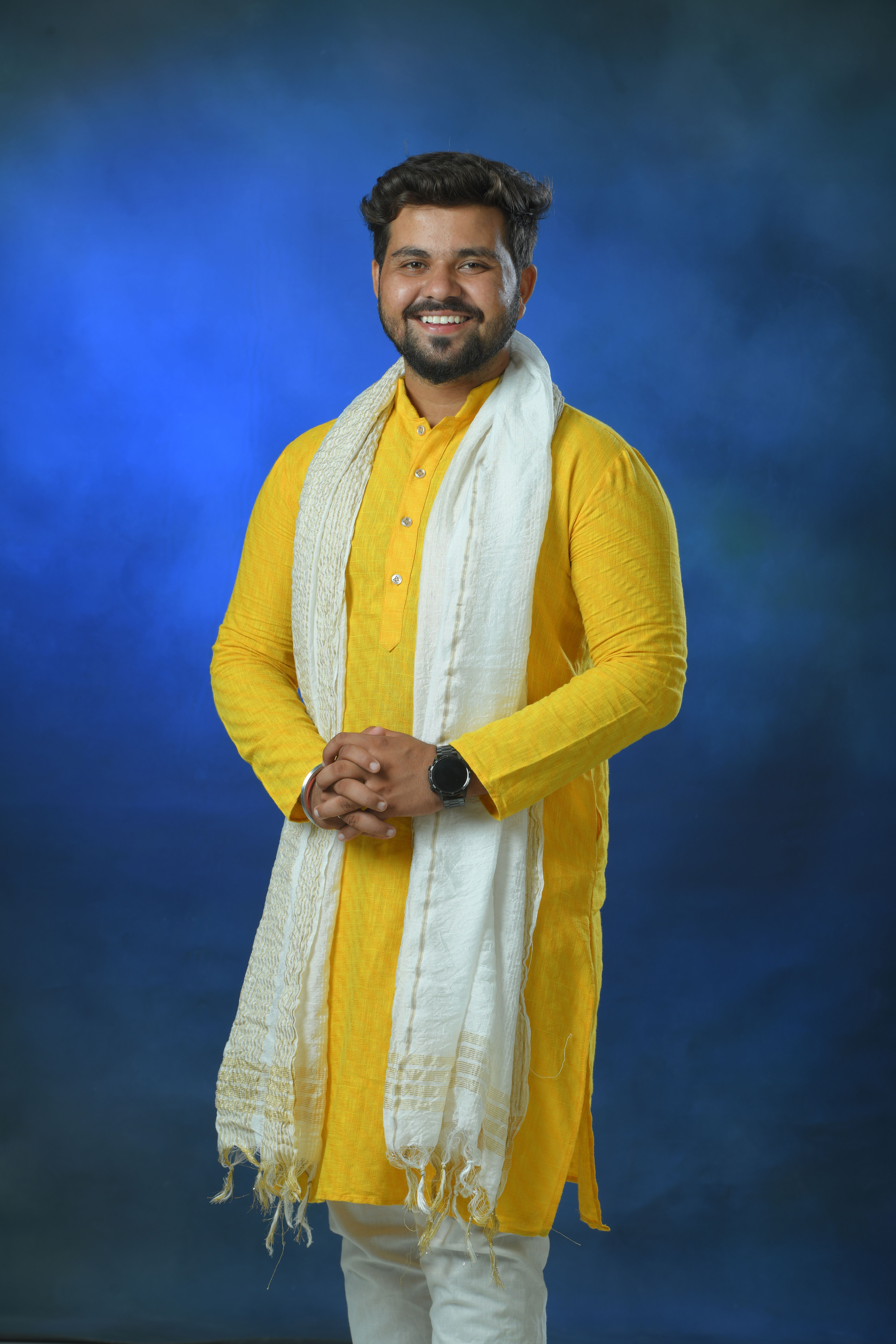 Portrait of a man in a yellow kurta and scarf, posing confidently against a blue background.