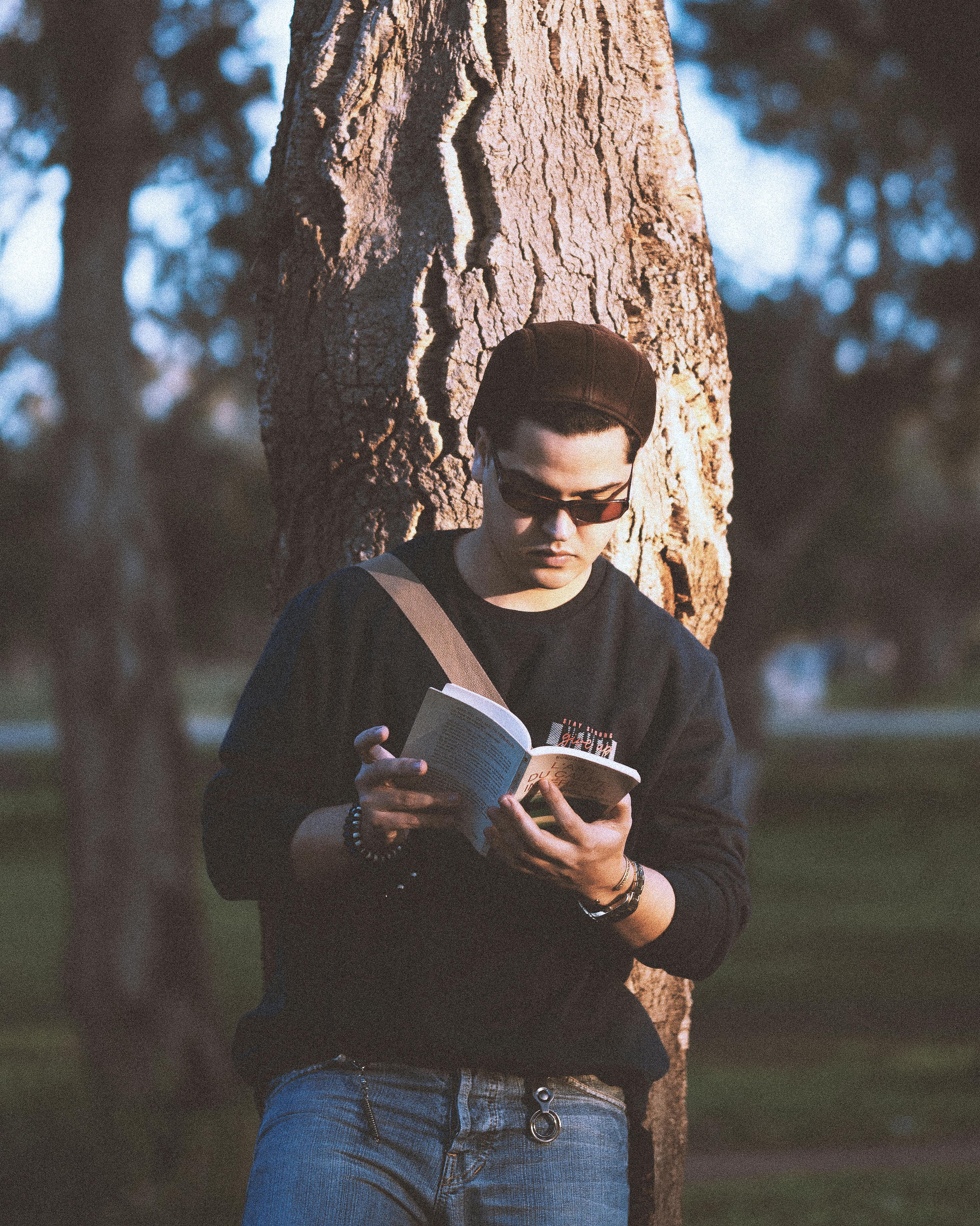 ingyenes Lazán öltözött fiatalember egy fának támaszkodva olvas egy könyvet a szabadban egy napsütéses napon. Stockfotó