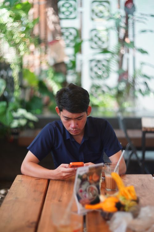 Free A young man engrossed in his phone, sitting in a cozy plant-filled café. Stock Photo