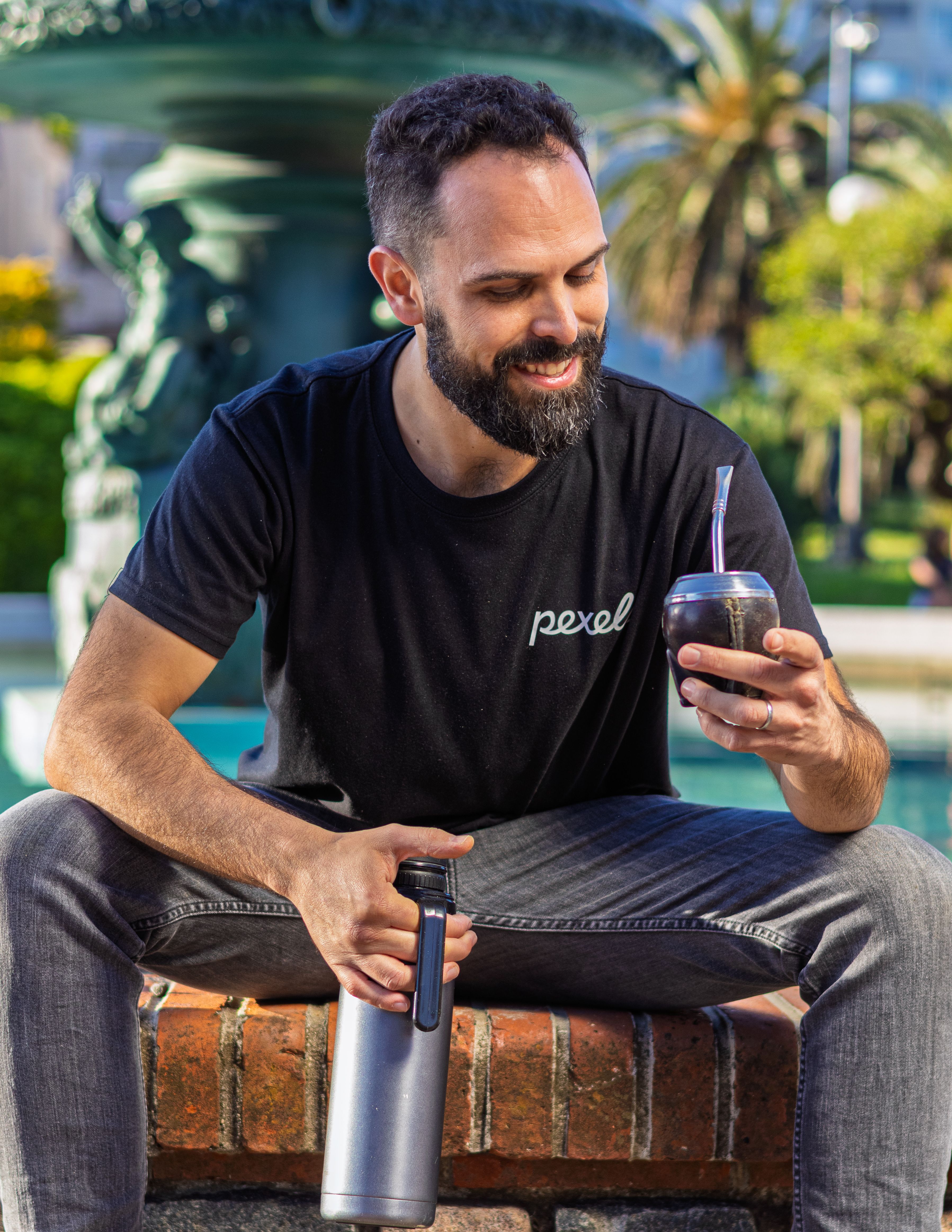 A man enjoys mate with a thermos in a bright urban park setting.