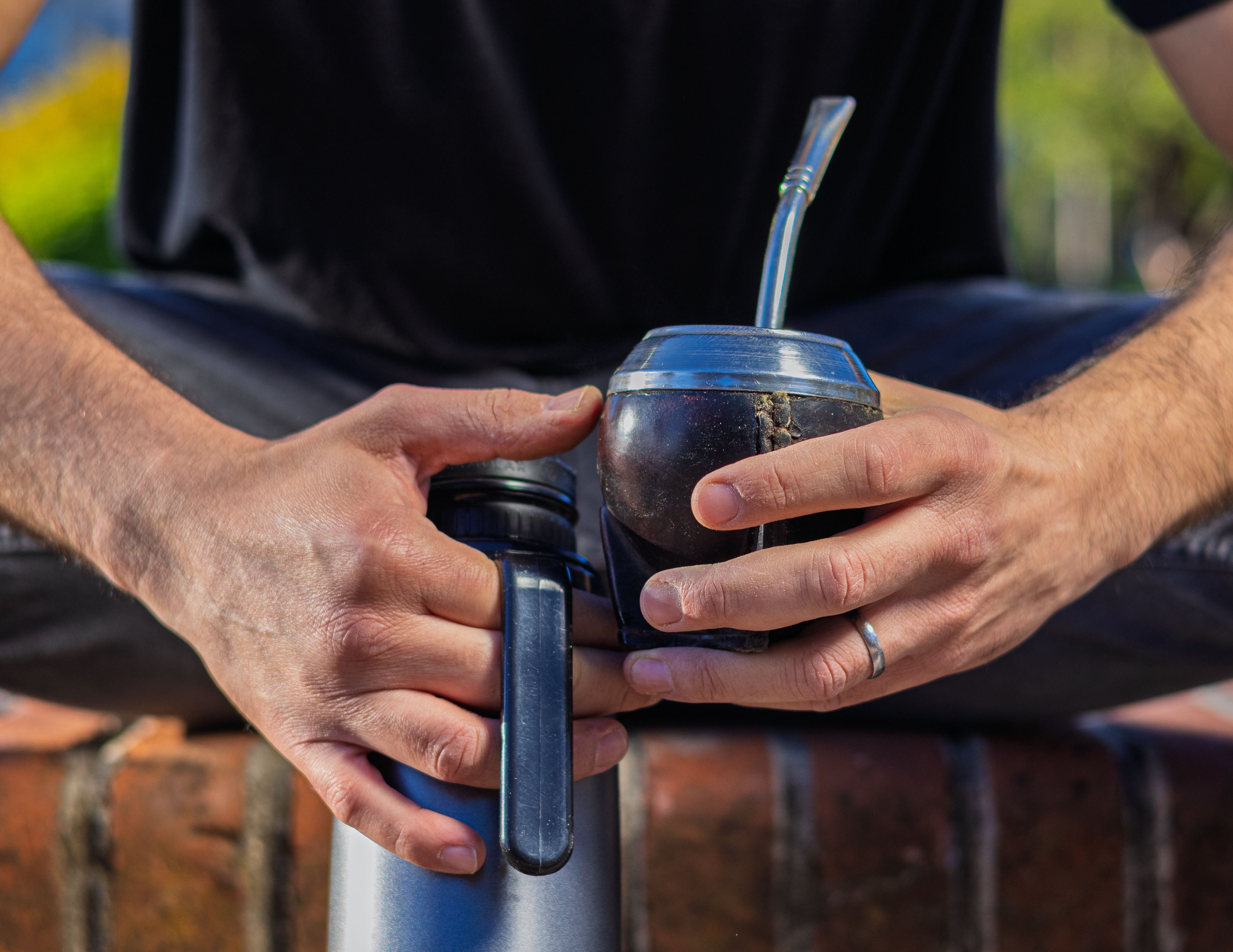 Close-up of man preparing yerba mate drink with thermos and cup in an outdoor setting.