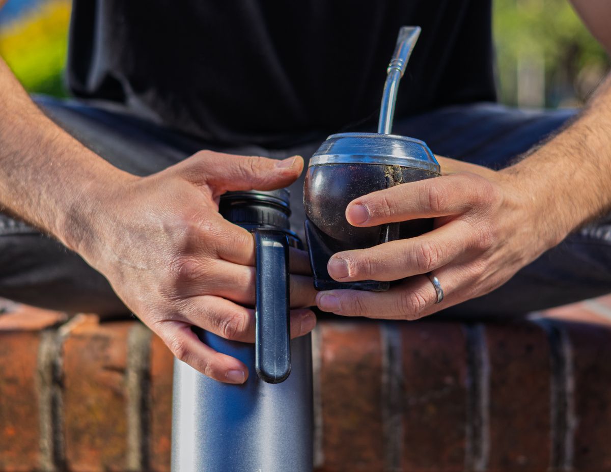 Close-up of a man's hands holding a mate gourd with its metal bombilla straw and a thermos, seated outdoors