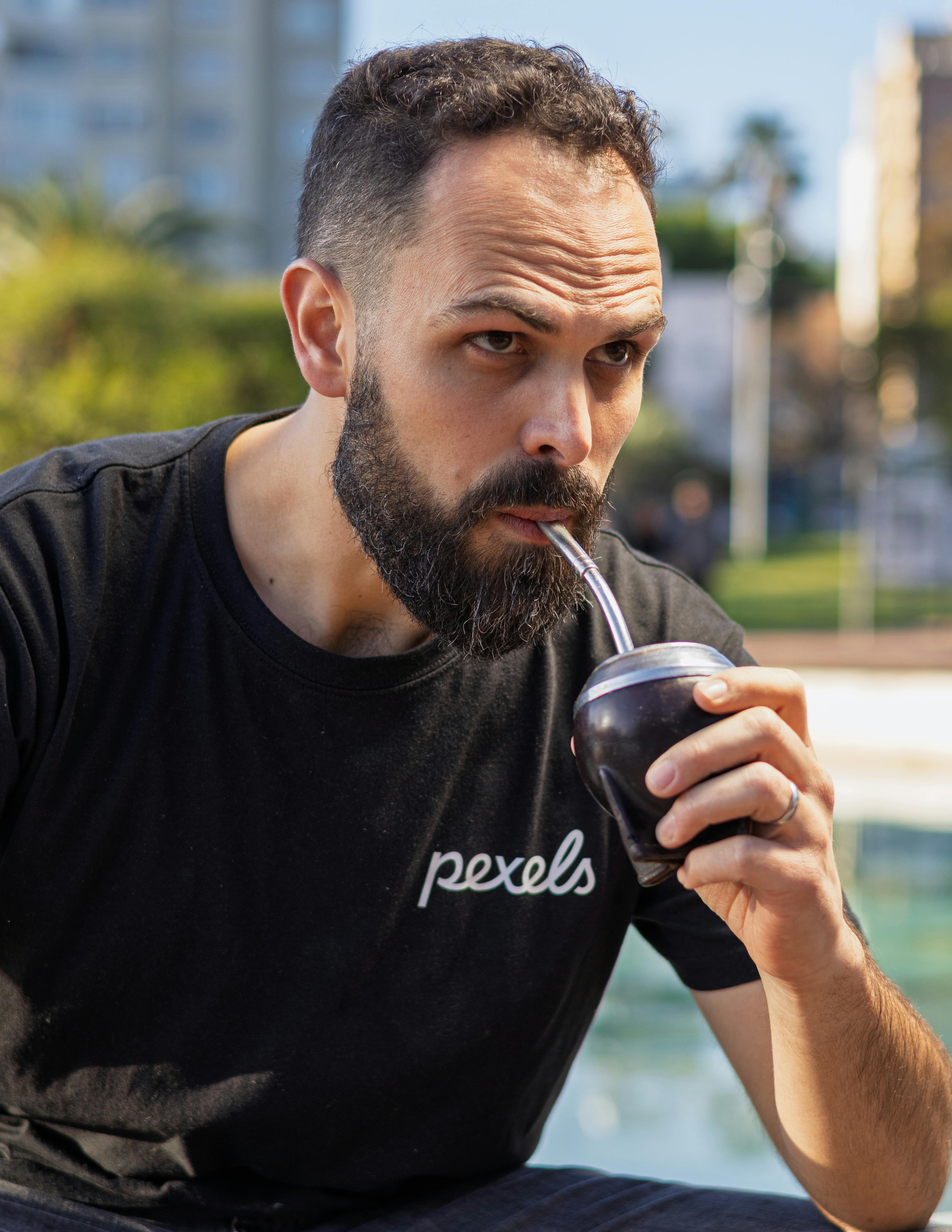 A bearded man enjoys yerba mate from a traditional gourd outdoors on a sunny day.