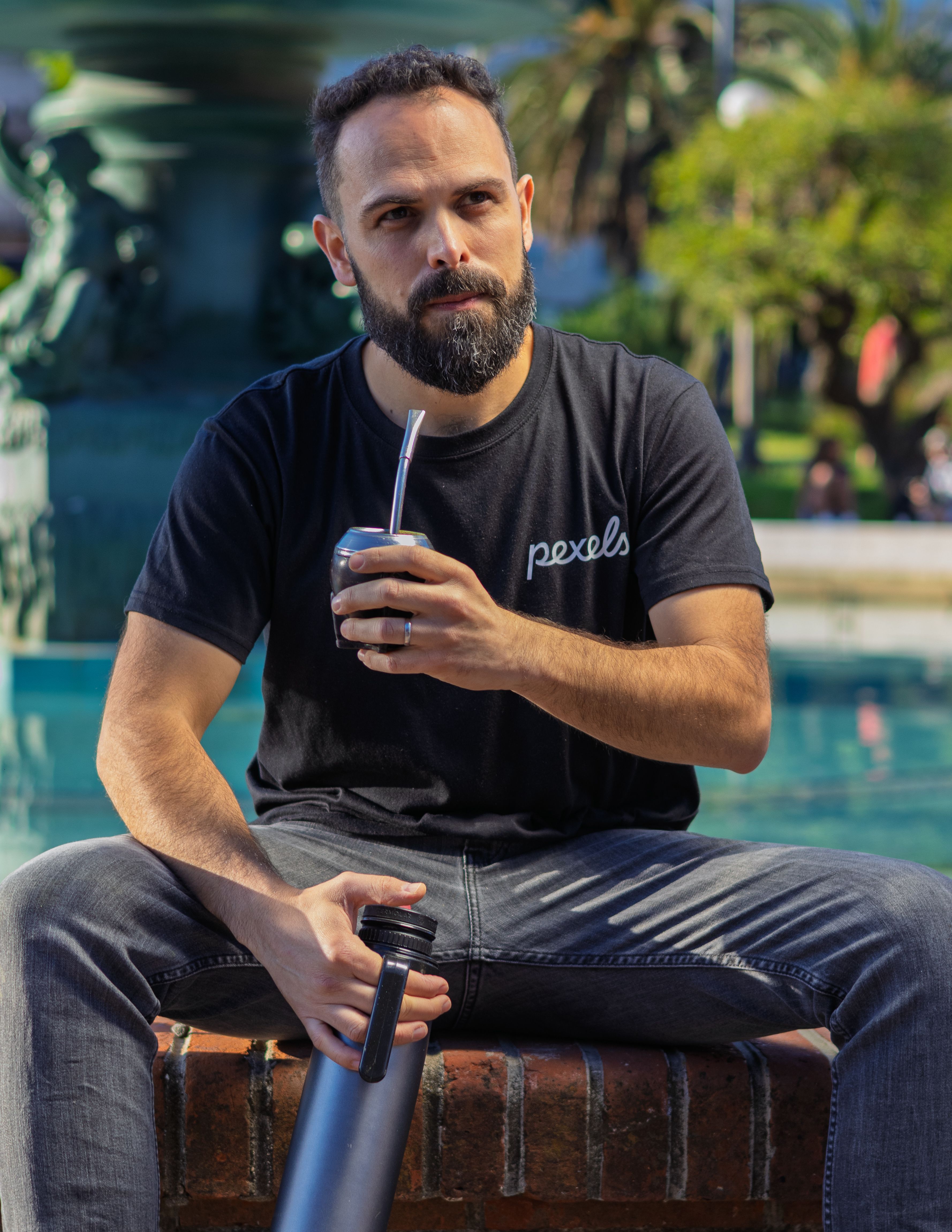 Bearded man sipping mate in sunny outdoor setting with fountain background.