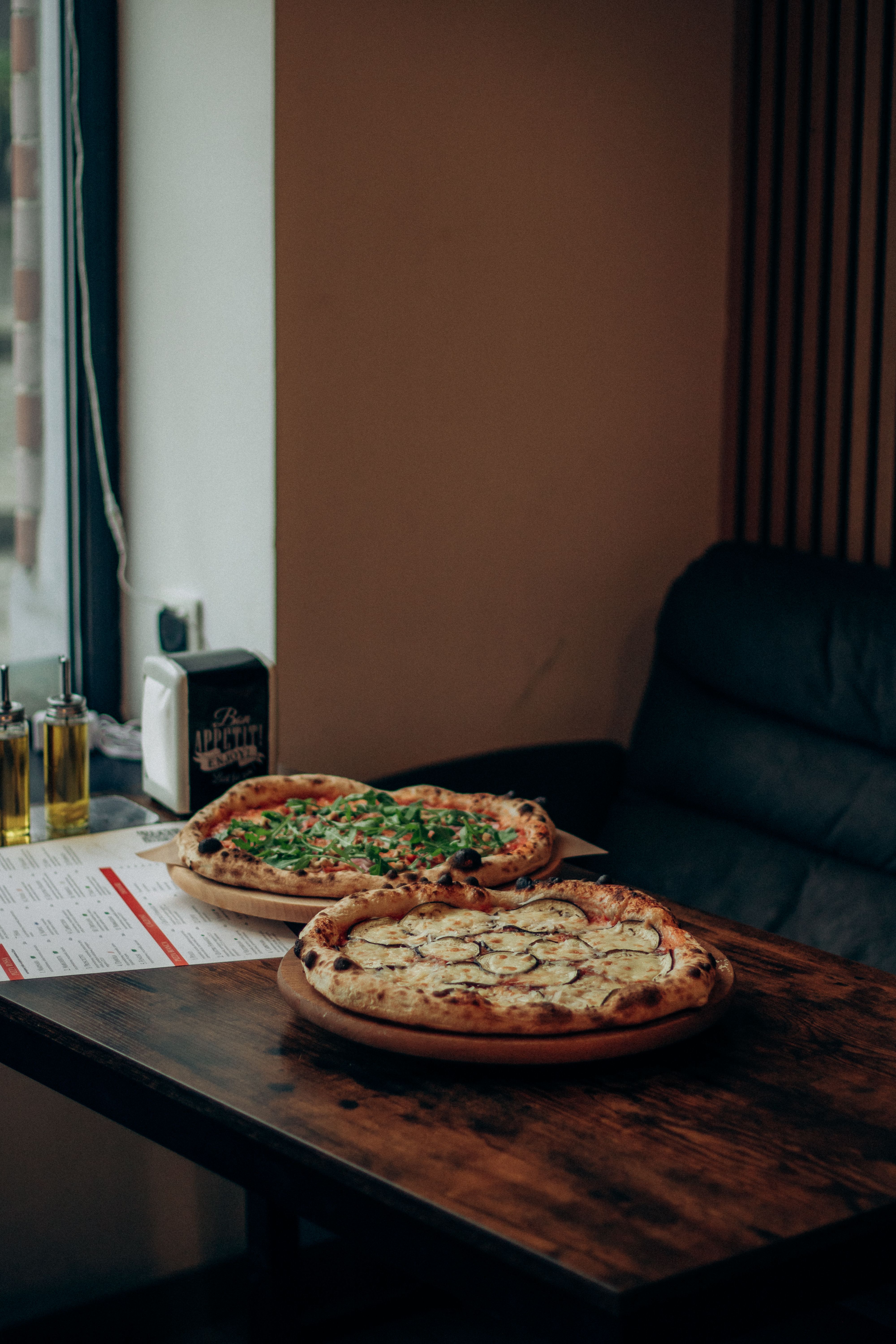 Two gourmet pizzas on a wooden table near a window in a cozy restaurant.