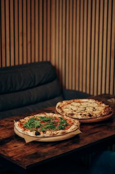 Two artisan pizzas with toppings on a rustic wooden table indoors.