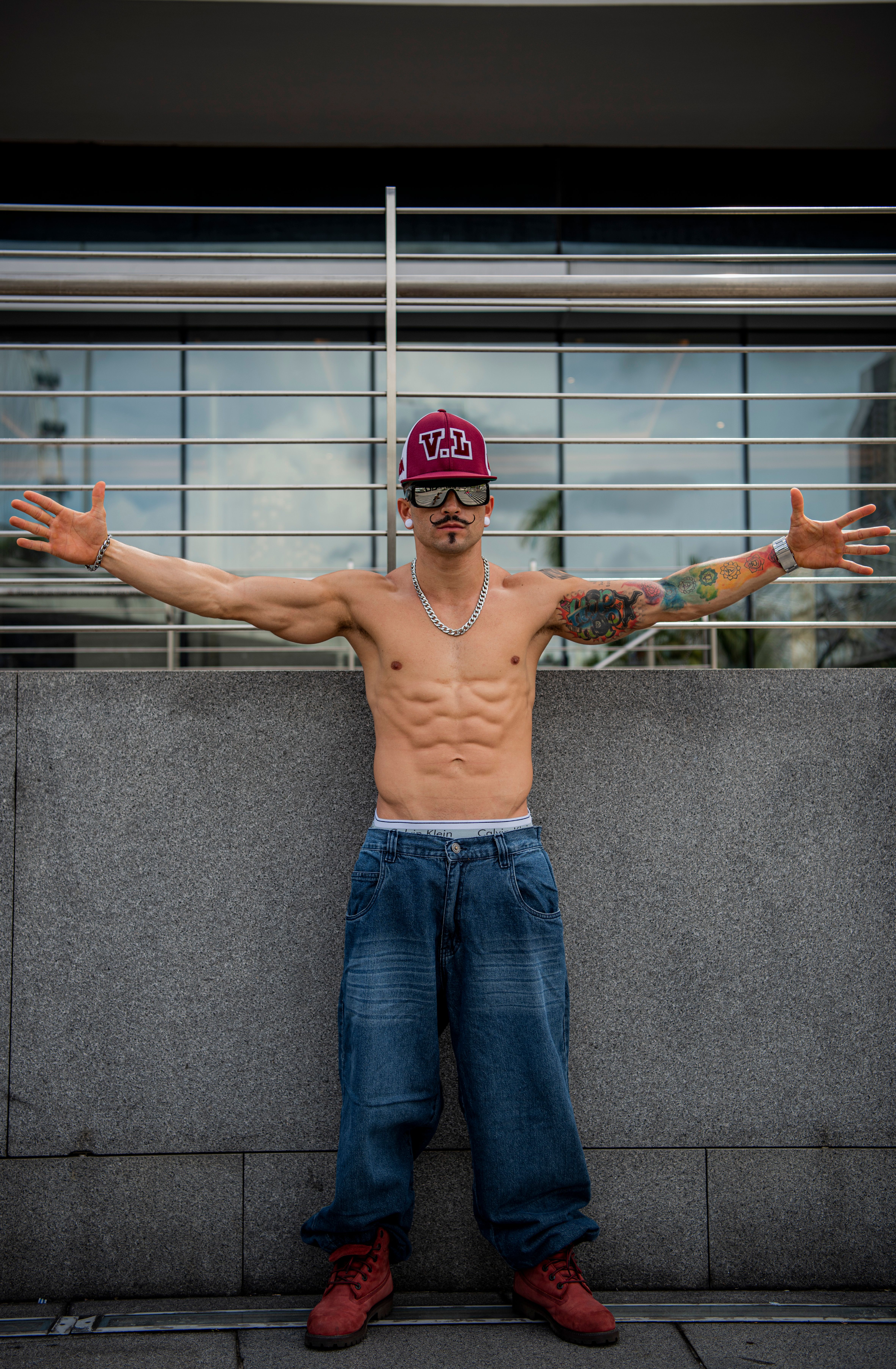 Tattooed shirtless man with cap and jeans posing confidently in an urban environment.