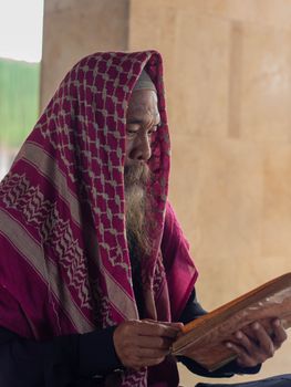 Elderly Middle Eastern man reads Quran peacefully indoors wearing a traditional keffiyeh.