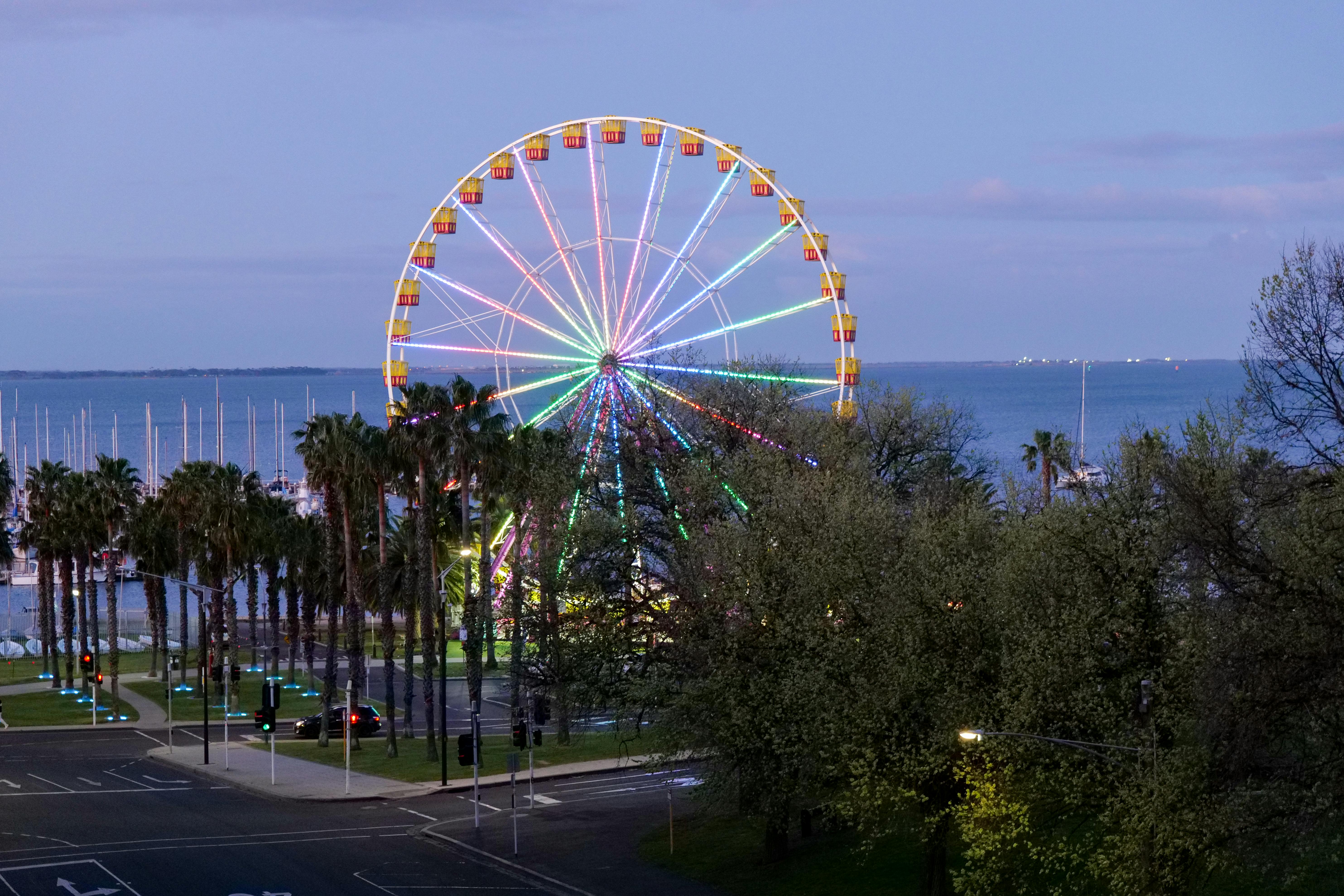 A vibrant Ferris wheel lights up the evening in Geelong, overlooking the serene bay and palm-lined streets.