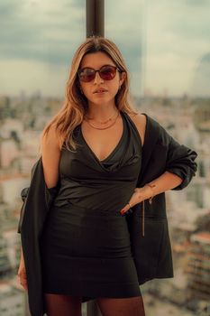 Stylish woman wearing sunglasses poses confidently in front of a cityscape. Urban fashion and empowerment.