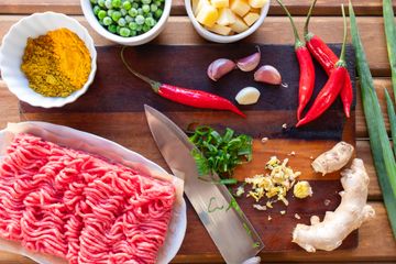 Fresh ground meat beside chilies, garlic, ginger, curry powder, peas and potatoes on a board