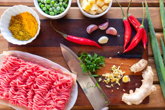 Fresh ground meat beside chilies, garlic, ginger, curry powder, peas and potatoes on a board