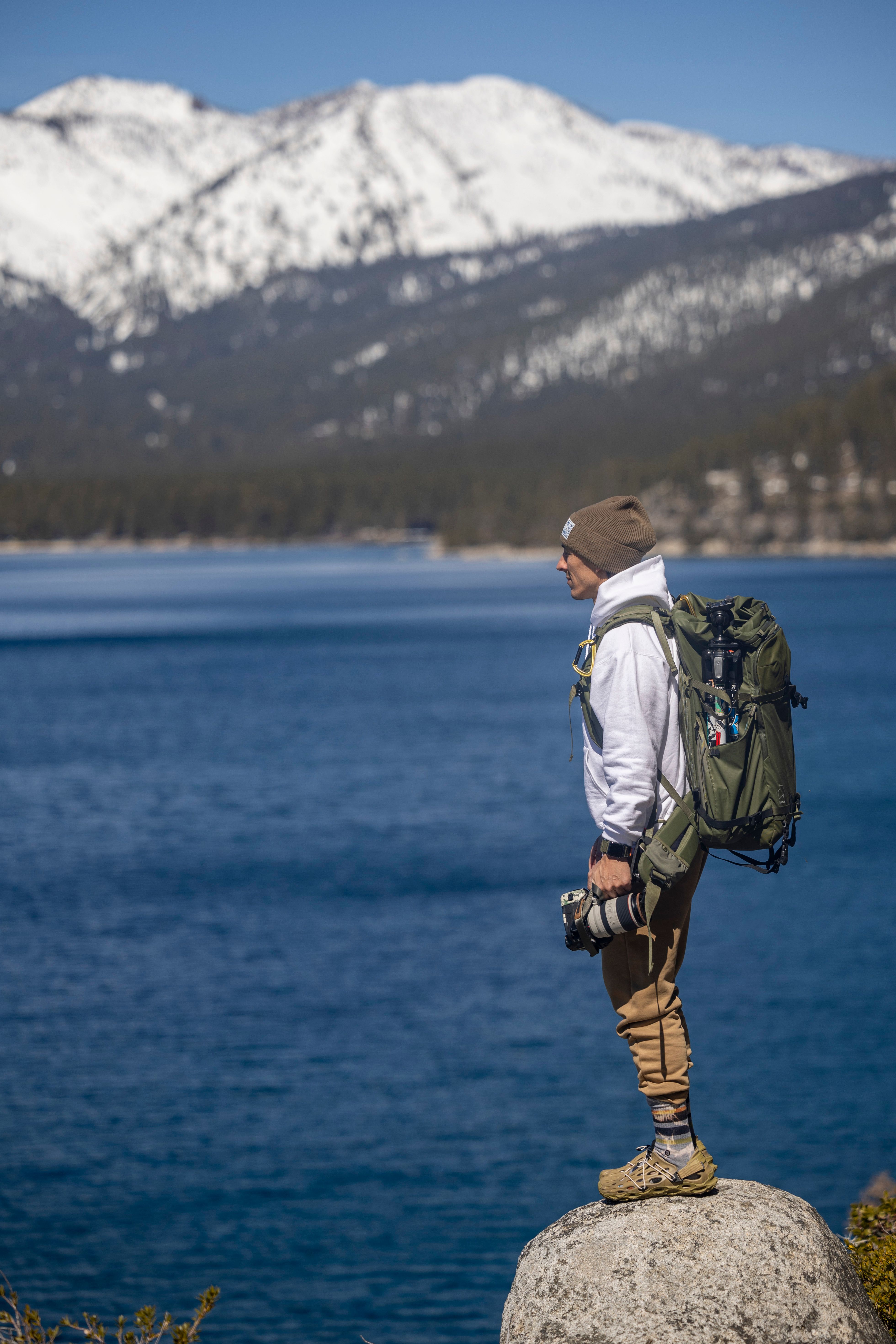 A photographer stands on a rock overlooking a tranquil lake with snow-capped mountains in the background.