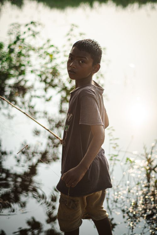 Free A young boy stands beside a serene lake at twilight, surrounded by nature. Stock Photo