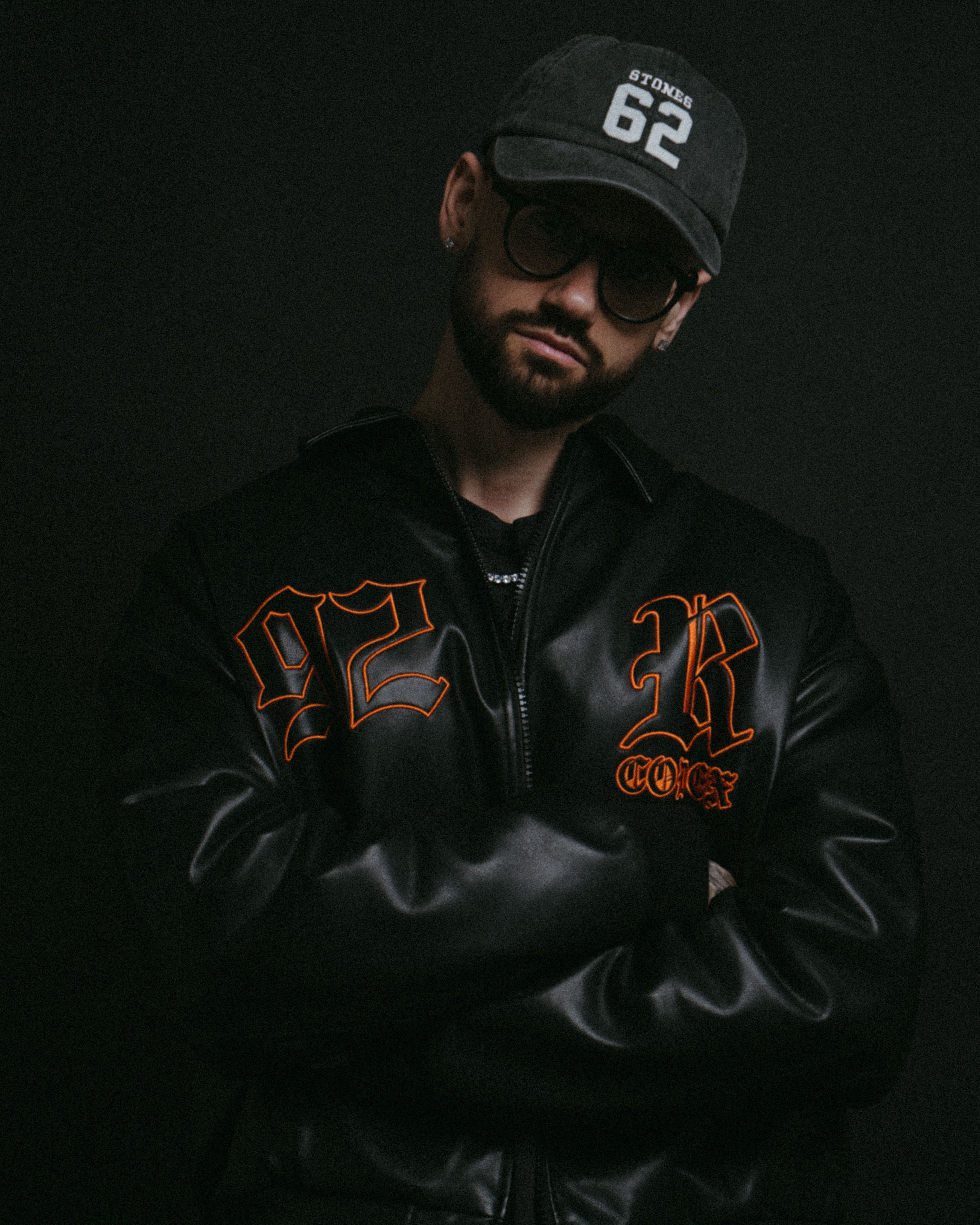 Portrait of a young man in dark clothing, wearing a cap and sunglasses, exuding urban fashion style.