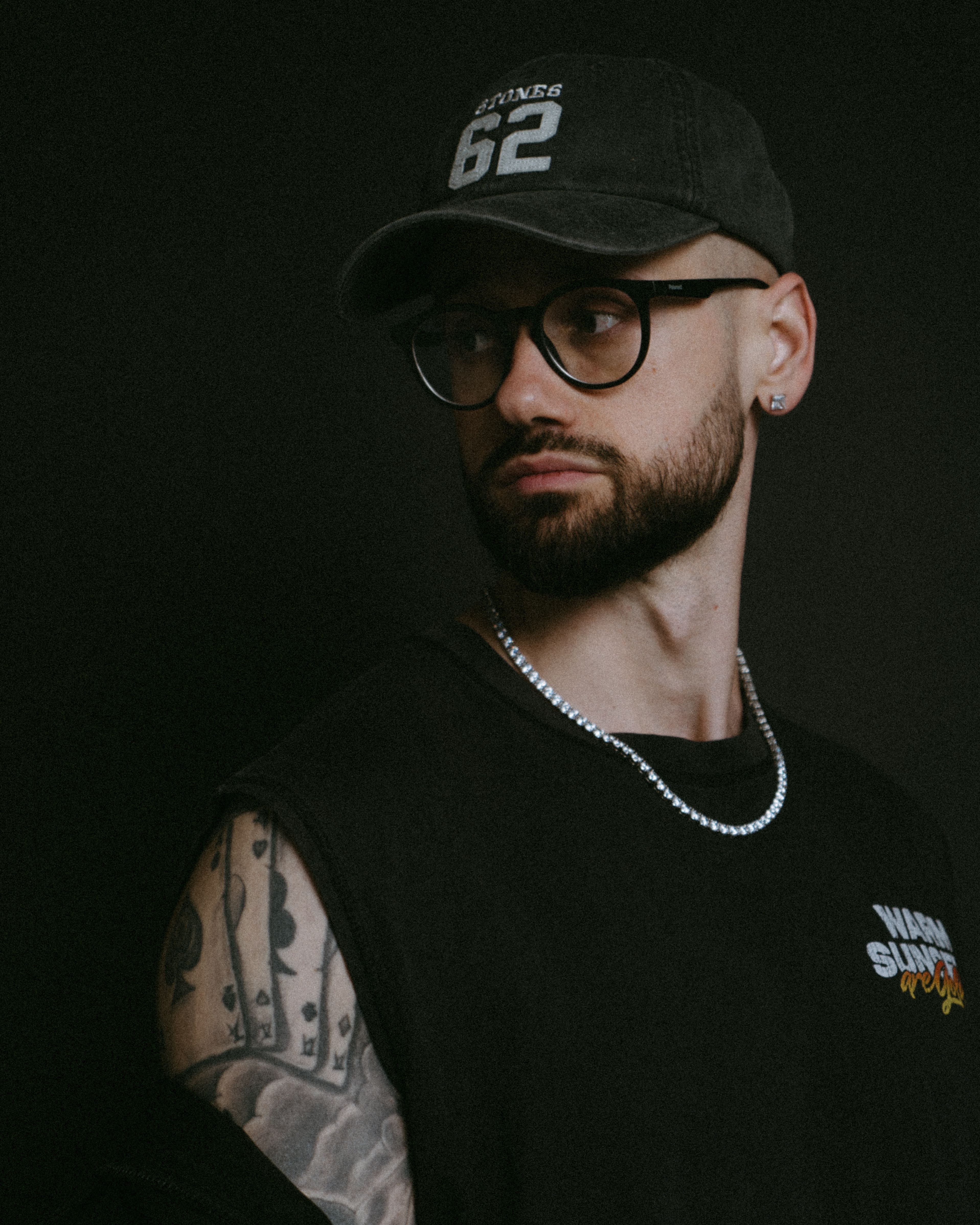 Portrait of a bearded man with tattoos and accessories against a dark background.