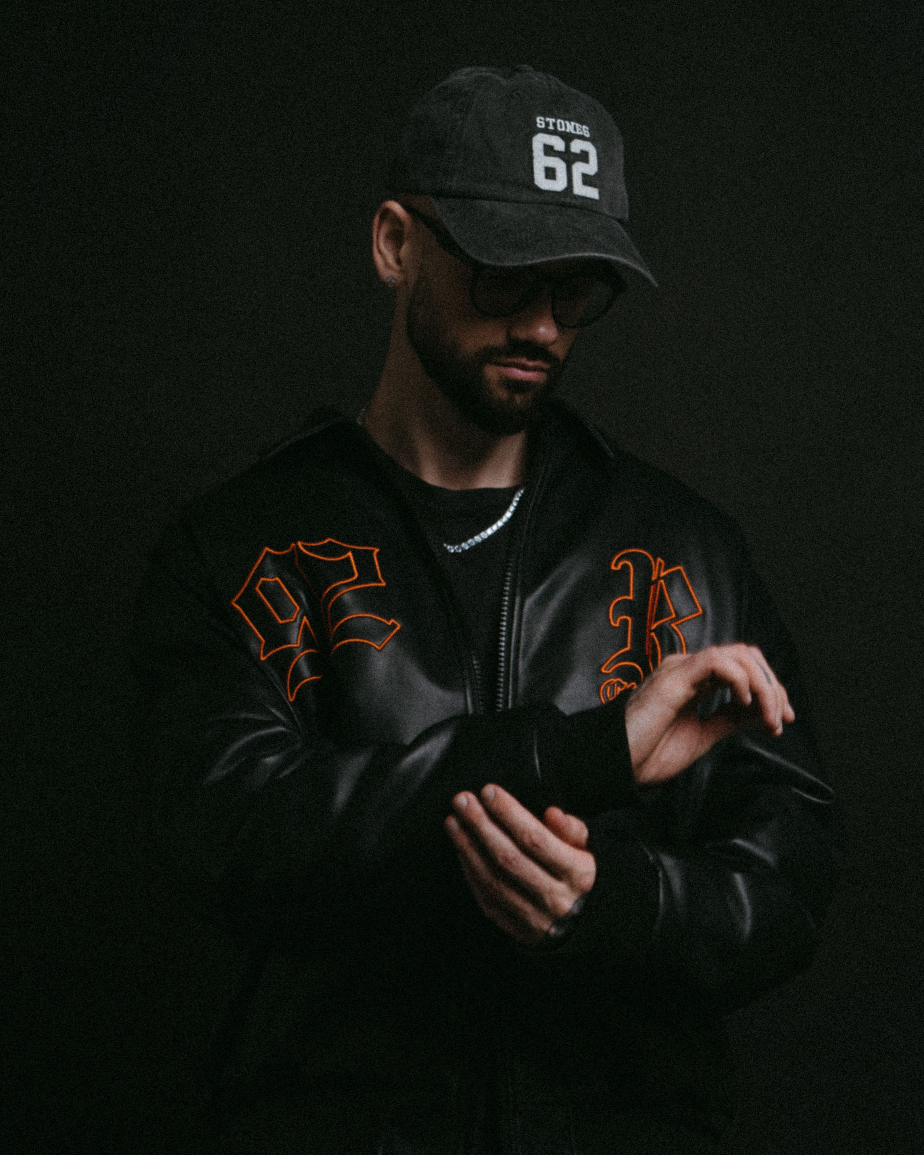 Man in dark jacket and cap in moody lighting, adjusting sleeve.