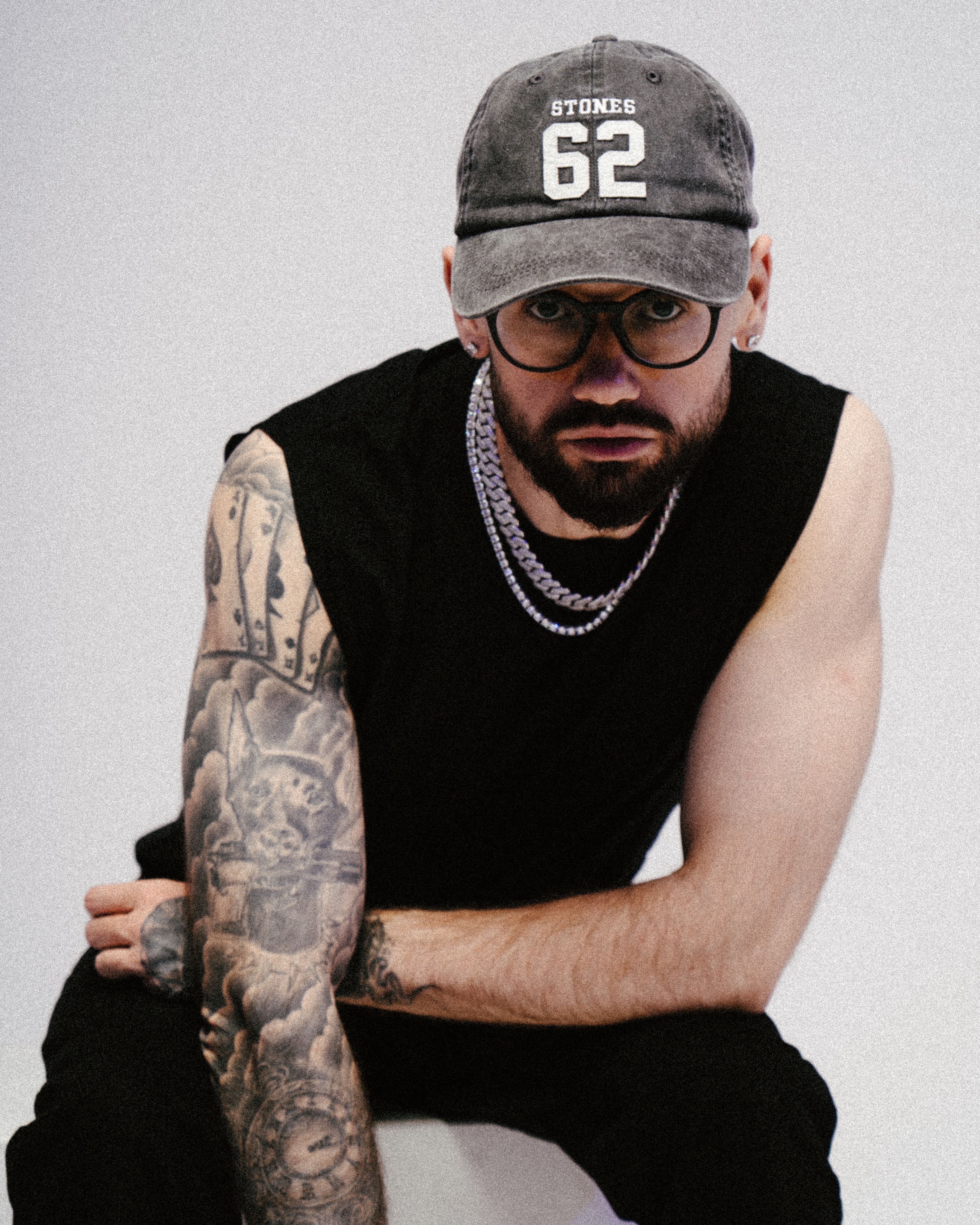 Portrait of a bearded man in casual attire, showcasing tattoos and wearing a cap in a studio setting.