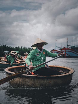 一名头戴圆锥帽的男子，在平静的河面上划着一艘传统竹船，周围还有其他划船的人