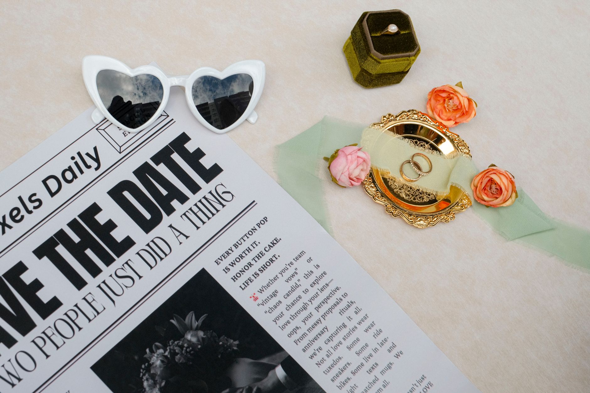 Charming flat lay of wedding essentials featuring rings, flowers, and sunglasses on a newspaper.